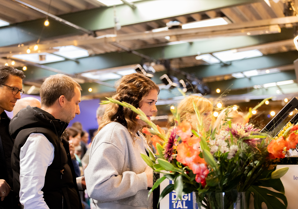 Das Bild zeigt eine Gruppe von Personen, die in einem Raum mit einem großen Blumenarrangement im Vordergrund stehen. Die Personen scheinen in ein Gespräch vertieft zu sein oder etwas im Raum zu beobachten. Der Raum hat eine grüne Decke, an der Lichterketten hängen, die eine warme und einladende Atmosphäre schaffen. Das Blumenarrangement steht auf einem Tisch und zieht die Aufmerksamkeit der Menschen im Raum auf sich.