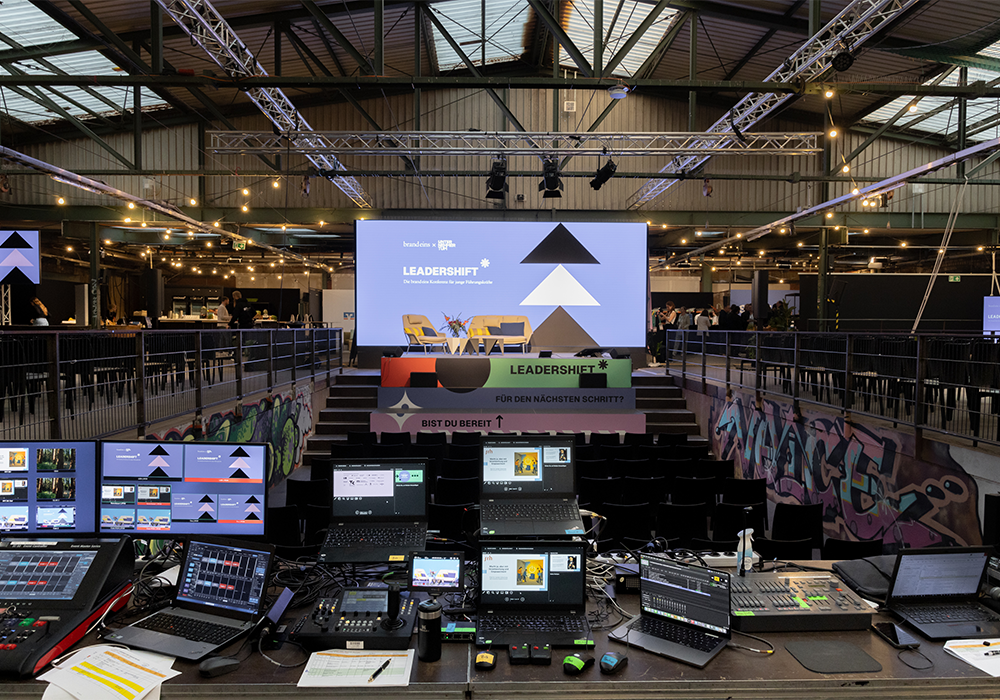 Das Bild zeigt einen großen Veranstaltungsraum mit einer Bühne, die für eine Präsentation aufgebaut ist. Die Bühne ist mit verschiedenen elektronischen Geräten ausgestattet, darunter Laptops, Monitore und Soundboards. Es sind auch mehrere Personen im Raum anwesend, die wahrscheinlich an der Veranstaltung teilnehmen oder die Einrichtung verwalten. Die Gesamtatmosphäre scheint professionell und gut organisiert zu sein, und die Bühne und die Ausrüstung sind für eine ansprechende Präsentation vorbereitet.