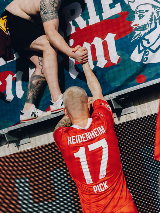 Das Bild zeigt eine Person in einem roten Trikot mit der Aufschrift "Heidenheim" und der Nummer "17", die einer anderen Person in einem schwarzen Hemd die Hand reicht. Beide stehen auf einer Plattform mit einem großen Transparent im Hintergrund.