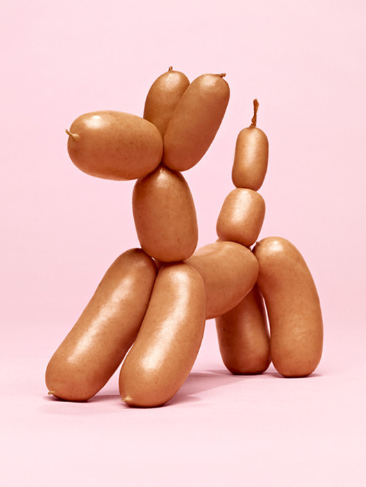 Das Bild zeigt eine kreative Anordnung von Hot Dogs auf einem rosa Hintergrund. Die Hot Dogs sind gestapelt und miteinander verbunden, um die Form eines Hundes zu bilden, wobei die Hot Dogs in Größe und Position variieren, um ein spielerisches und visuell ansprechendes Design zu schaffen.