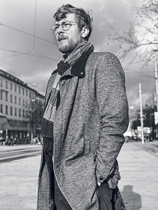 Ein Mann in einem grauen Mantel und mit Brille steht auf einer Straße in der Stadt, den Blick zur Seite gerichtet. Er trägt einen Schal um den Hals und scheint etwas oder jemanden außerhalb des Bildausschnitts zu betrachten. Das Bild ist in Schwarz-Weiß gehalten, was ihm eine zeitlose Qualität verleiht. Der Mann ist im Vordergrund positioniert, während die Stadtlandschaft und die Gebäude den Hintergrund bilden.