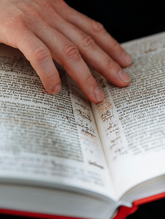 Das Bild zeigt eine Nahaufnahme der Hand einer Person, die auf einem offenen Buch mit handschriftlichen Notizen ruht. Die Finger der Person sind sichtbar, und das Buch scheint einen roten Einband zu haben. Die handschriftlichen Notizen sind über die Seiten verstreut, was darauf hindeutet, dass die Person mit dem Lesen oder Studieren beschäftigt ist. Das Bild enthält keinen erkennbaren Text, und der Schwerpunkt liegt auf der Interaktion zwischen der Hand und dem Buch.