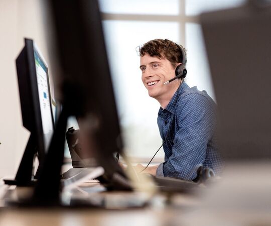 Ein lächelnder Mann, der ein Headset trägt, sitzt an einem Schreibtisch und arbeitet mit einem Headset an einem Computer. Der Schreibtisch ist mit einem Monitor, einer Tastatur und einer Maus ausgestattet, und auf dem Schreibtisch befindet sich auch ein Laptop. Der Hintergrund ist unscharf und lenkt die Aufmerksamkeit auf den Mann und seinen Arbeitsbereich.
