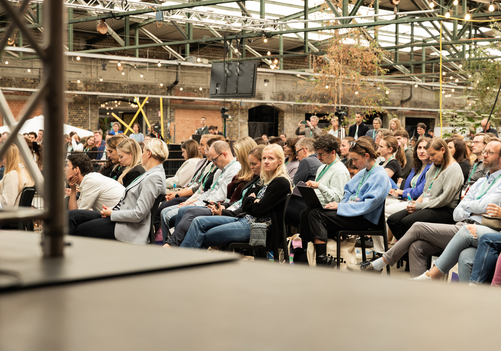 Das Bild zeigt eine große Gruppe von Menschen, die auf Stühlen sitzen und wahrscheinlich an einer Veranstaltung oder Konferenz teilnehmen. Die Personen sind zur Vorderseite des Raums gerichtet, was darauf hindeutet, dass sie einem Redner oder einer Präsentation zuhören. Die Umgebung scheint ein überdachter Veranstaltungsort mit einer hohen Decke und freiliegenden Balken zu sein, was einen rustikalen oder industriellen Eindruck vermittelt. Das Bild enthält keinen erkennbaren Text, und die relativen Positionen der Personen und Stühle deuten auf eine typische Sitzordnung für solche Versammlungen hin.