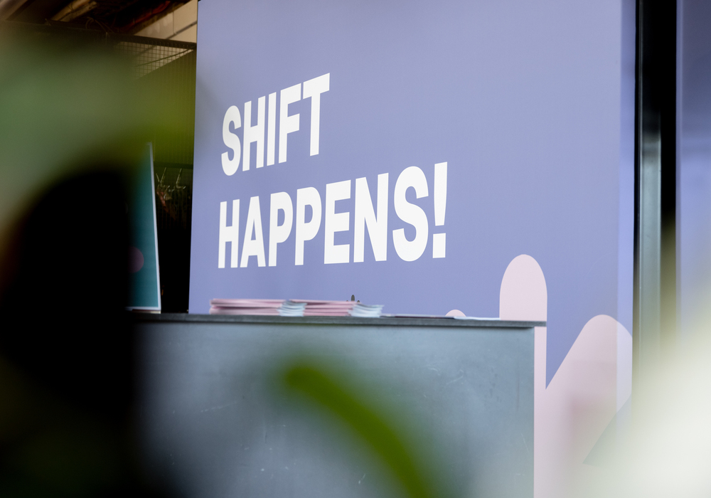 Das Bild zeigt ein großes, lilafarbenes Schild mit der Aufschrift "SHIFT HAPPENS!" in weißen Buchstaben. Das Schild befindet sich in einem Innenraum mit einem unscharfen Hintergrund, der eine Pflanze und eine Theke enthält. Das Schild scheint Teil einer größeren Ausstellung oder Installation zu sein, möglicherweise im Zusammenhang mit einem Geschäft oder einer Veranstaltung. Das Bild enthält keinen identifizierbaren Text oder identifizierbare Objekte, und die Gesamtkomposition legt nahe, dass der Schwerpunkt auf der auffälligen Botschaft auf dem Schild liegt.