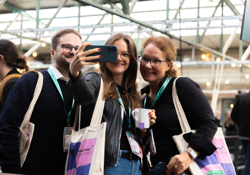 Drei Personen stehen zusammen, lächeln und machen ein Selfie mit einem Smartphone. Sie tragen Namensschilder und haben Taschen dabei, was darauf hindeutet, dass sie an einer Veranstaltung oder Konferenz teilnehmen könnten. Der Hintergrund scheint ein großer offener Raum mit einigen Lichtern zu sein, was darauf hindeutet, dass das Foto in einem gut beleuchteten Bereich aufgenommen wurde.