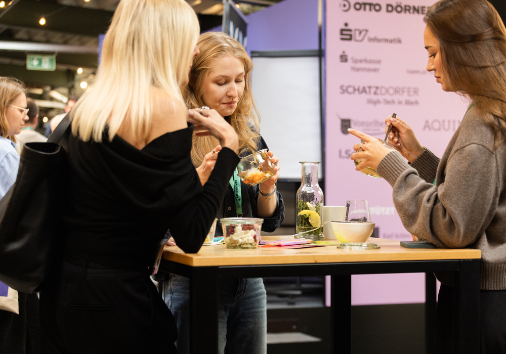 Das Bild zeigt drei Frauen, die um einen Tisch herum stehen, sich unterhalten und Essen und Getränke genießen. Der Tisch ist mit verschiedenen Gegenständen gedeckt, darunter eine Schüssel, eine Tasse und eine Flasche. Im Hintergrund ist ein Schild mit Text und Logos zu sehen, was auf ein gesellschaftliches oder berufliches Treffen hindeutet. Die Frauen scheinen sich auf ihr Gespräch zu konzentrieren, wodurch eine lebhafte Atmosphäre entsteht.