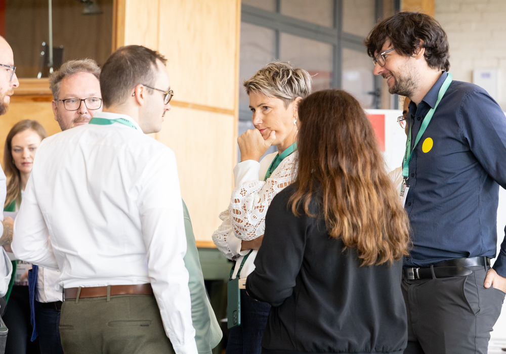 Das Bild zeigt eine Gruppe von Personen, die in eine Diskussion vertieft sind, wobei einige stehen und andere um einen Tisch sitzen. Die Personen scheinen sich in einem professionellen Umfeld zu befinden, möglicherweise in einer Konferenz oder Besprechung. Der Raum hat einen Holzboden und eine Glaswand, was eine warme und einladende Atmosphäre schafft. Die Personen tragen Geschäftskleidung, was auf eine formelle oder professionelle Veranstaltung hindeutet.