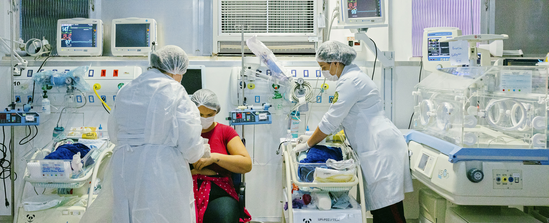 Das Bild zeigt ein Krankenhauszimmer mit medizinischen Geräten und Personal. Es sind zwei Personen in weißen Kitteln zu sehen, eine sitzend und eine stehend, beide tragen Masken. Der Raum ist mit Monitoren und anderen medizinischen Geräten ausgestattet. Ein Baby wird von einer Krankenschwester betreut, die die Hand des Babys hält. 