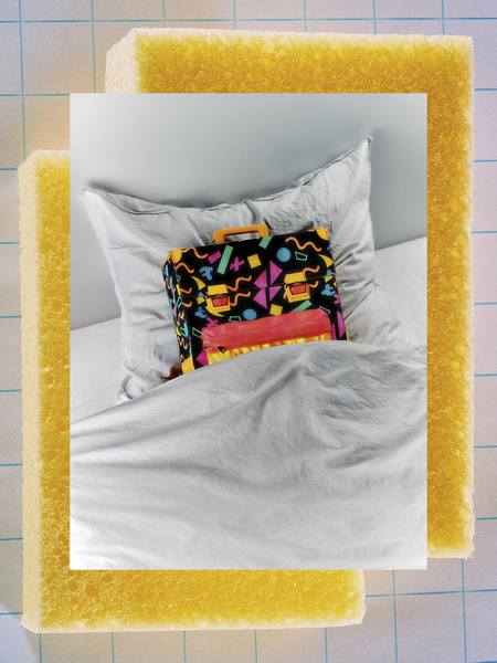 Das Bild zeigt einen farbenfrohen, gemusterten Koffer, der auf einem Bett mit einem weißen Kissen steht. Der Koffer befindet sich in der Mitte des Bettes, das Kissen liegt darauf. Der Hintergrund des Bildes ist ein Gitter aus gelben Schaumstoffblöcken, das einen interessanten Kontrast zu dem bunten Koffer bildet. Die Gesamtkomposition des Bildes lässt auf eine kreative oder künstlerische Gestaltung schließen, möglicherweise für eine Website oder Werbung.