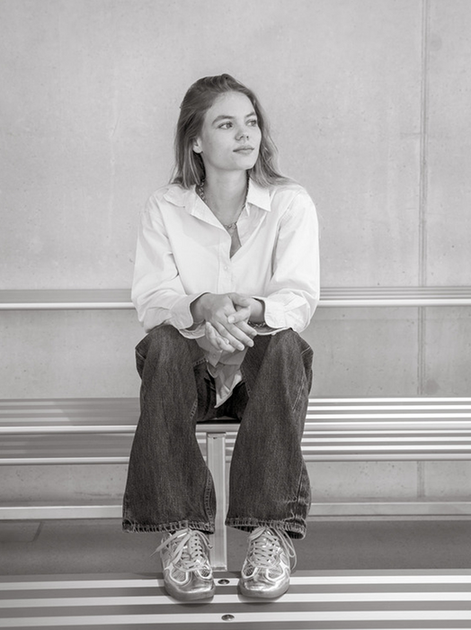 Eine junge Frau sitzt auf einer Bank und blickt mit vor sich verschränkten Händen zur Seite. Sie trägt ein weißes Hemd, Jeans und Turnschuhe. Das Bild ist in Schwarz-Weiß gehalten, was ihm eine zeitlose Qualität verleiht. Die Frau ist in der Mitte des Bildes positioniert und nimmt den größten Teil des Bildes ein. Die Bank, auf der sie sitzt, befindet sich im Vordergrund, während der Hintergrund eine einfache Wand ist.