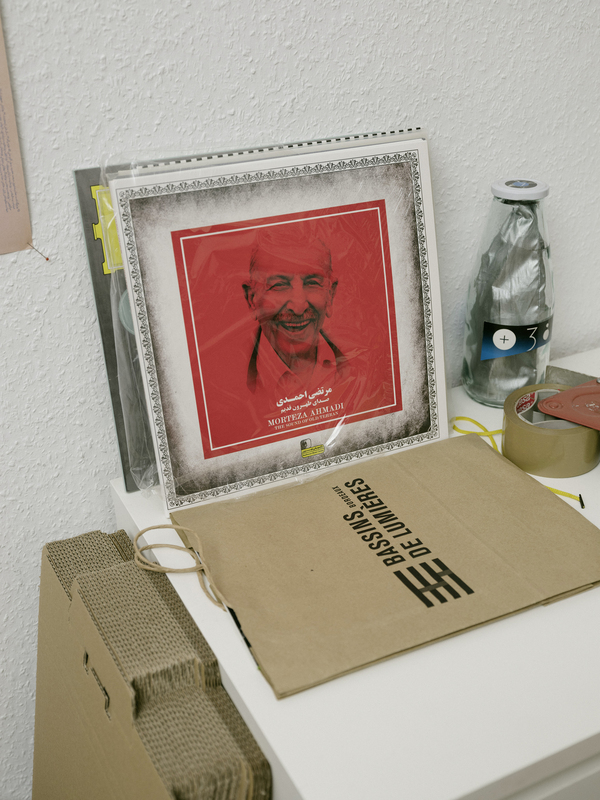 Das Bild zeigt ein gerahmtes Foto eines Mannes mit rotem Hintergrund, das auf einer weißen Fläche steht. Neben dem gerahmten Foto befindet sich ein Karton mit Text, und im Hintergrund ist eine Flasche zu sehen. Die gesamte Szene scheint eine einfache Anordnung von Alltagsgegenständen zu sein.