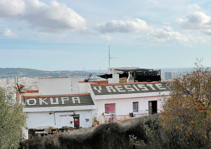 Das Bild zeigt ein weißes Gebäude mit einem schwarzen Dach, das auf einem Hügel mit Blick auf eine Stadtlandschaft steht. Auf dem Dach des Gebäudes sind die Worte "OKUFA" und "RESIST" zu lesen. Die Szene befindet sich vor einem teilweise bewölkten Himmel im Hintergrund.