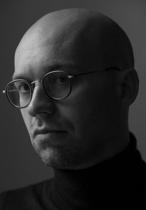 Das Bild ist ein Schwarz-Weiß-Porträt eines Mannes mit Glatze, der eine runde Brille und einen Rollkragenpullover trägt. Er blickt zur Seite und hat einen ernsten Gesichtsausdruck. Das Bild ist im Hochformat gehalten, und der Mann ist die zentrale Figur im Bild.