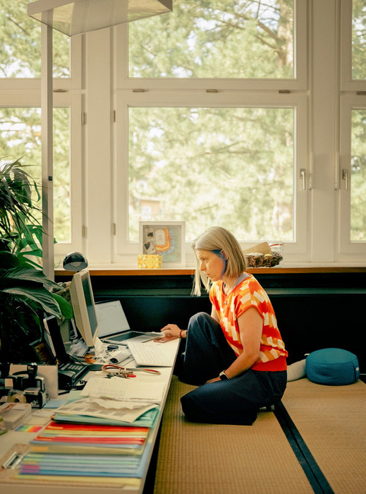 Eine Frau sitzt auf dem Boden vor einem Schreibtisch und arbeitet an einem Laptop-Computer. Der Schreibtisch ist mit verschiedenen Gegenständen vollgestopft, darunter eine Tastatur, eine Maus und mehrere Bücher. Der Raum hat große Fenster, die viel Tageslicht hereinlassen, und in der Nähe steht eine Topfpflanze. Die Frau scheint sich auf ihre Arbeit zu konzentrieren, möglicherweise mit Aufgaben im Zusammenhang mit ihrer Arbeit oder ihrem Studium.