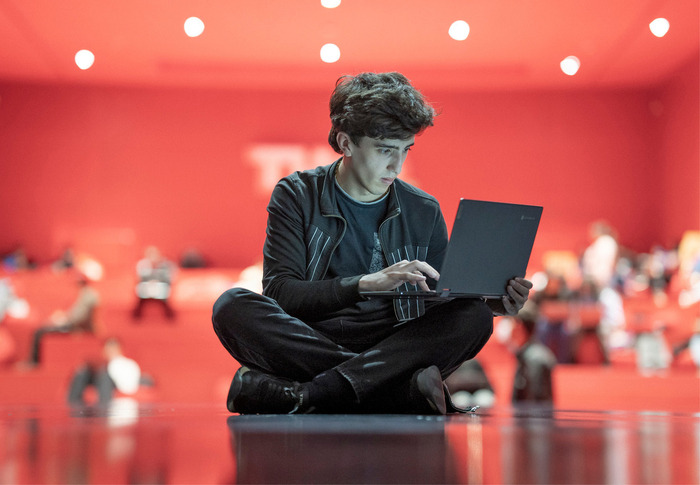 Das Bild zeigt einen jungen Mann, der im Schneidersitz auf dem Boden sitzt und sich auf die Arbeit mit einem Laptop konzentriert. Der Hintergrund ist unscharf und deutet auf einen großen Raum oder ein Auditorium hin. Der Mann scheint das Hauptmotiv des Bildes zu sein, wobei der Laptop das zentrale Objekt ist. Das Bild enthält keinen erkennbaren Text, und die Position des Mannes und die Hintergrundelemente erzeugen ein Gefühl von Tiefe und Konzentration auf den Laptop.