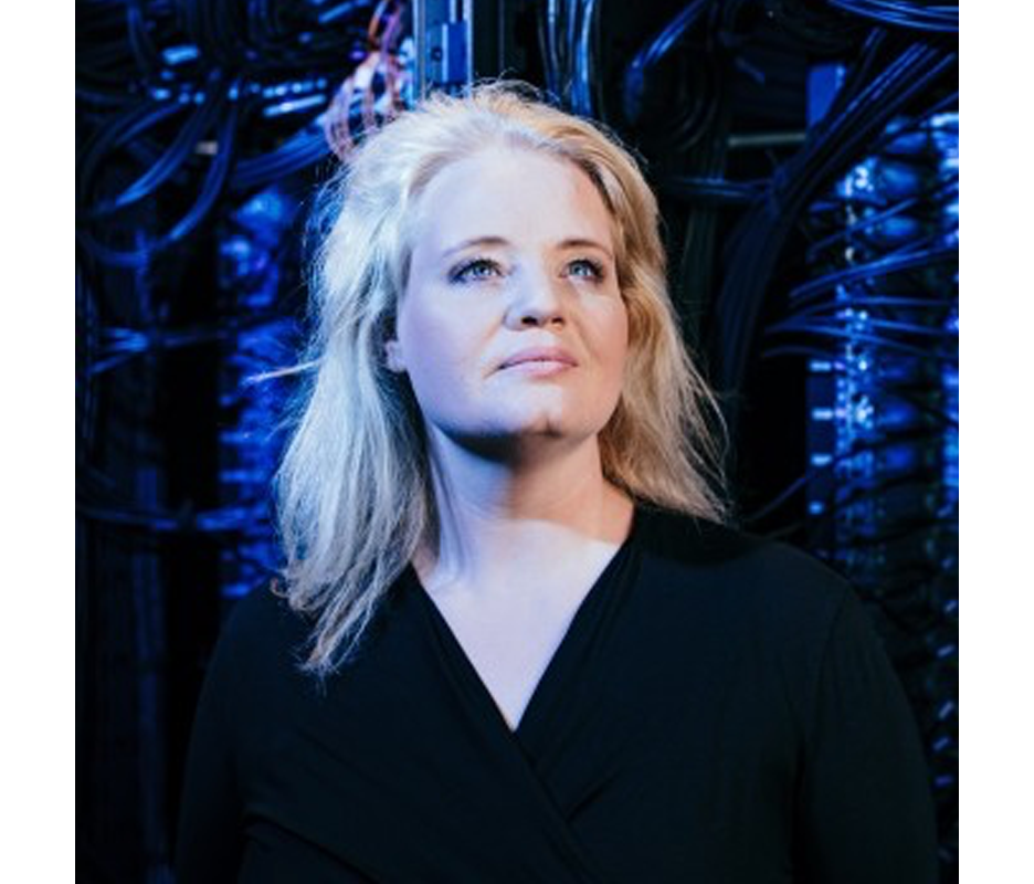 Das Bild zeigt eine Frau mit blondem Haar, die ein schwarzes Oberteil trägt und vor einem blauen Hintergrund mit verschiedenen Kabeln und Drähten steht. Die Frau scheint zur Seite zu blicken, möglicherweise um etwas oder jemanden zu beobachten. Das Bild enthält keinen erkennbaren Text, und die Position der Frau und die Hintergrundelemente lassen auf ein professionelles oder technisches Umfeld schließen.