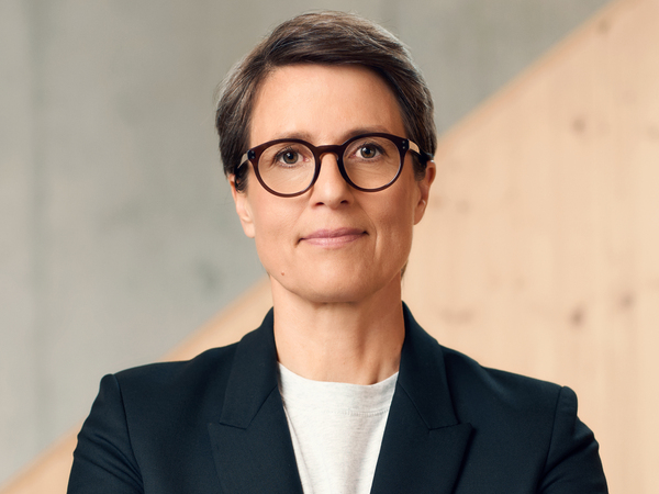 Das Bild zeigt eine Frau mit kurzen Haaren und Brille, die einen schwarzen Blazer und ein weißes T-Shirt trägt. Sie blickt direkt in die Kamera und hat einen neutralen Ausdruck. Der Hintergrund ist eine einfache graue Wand, die im Kontrast zur dunklen Kleidung der Frau steht. Das Bild ist ein Porträt, das das Gesicht und den Oberkörper der Frau zeigt.