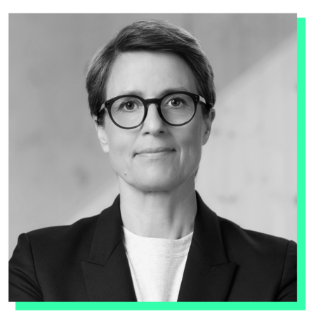 Das Bild ist ein Schwarz-Weiß-Porträt einer Frau, die eine Brille und einen Blazer trägt. Sie blickt mit neutralem Gesichtsausdruck direkt in die Kamera. Der Hintergrund ist eine schlichte weiße Wand, die einen Kontrast zu der Kleidung der Frau und der Schwarz-Weiß-Farbgebung des Bildes bildet. Das Bild enthält weder Text noch zusätzliche Objekte, und die Frau steht im Mittelpunkt der Komposition.