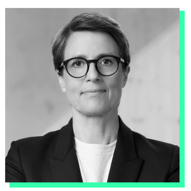 Das Bild ist ein Schwarz-Weiß-Porträt einer Frau, die eine Brille und einen Blazer trägt. Sie blickt mit neutralem Gesichtsausdruck direkt in die Kamera. Der Hintergrund ist eine schlichte weiße Wand, die einen Kontrast zu der Kleidung der Frau und der Schwarz-Weiß-Farbgebung des Bildes bildet. Das Bild enthält weder Text noch zusätzliche Objekte, und die Frau steht im Mittelpunkt der Komposition.