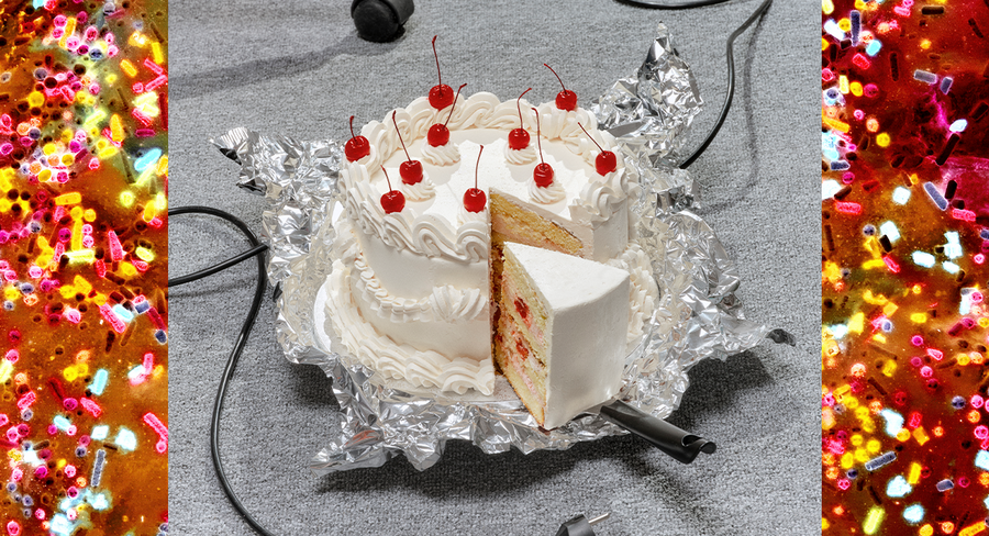 Das Bild zeigt einen weißen Kuchen mit roten Kirschen darauf, der auf einem Stück Alufolie steht. Ein Stück wurde vom Kuchen abgeschnitten, sodass man die Schichten und die Füllung sehen kann. Der Kuchen steht auf einer grauen Fläche, im Hintergrund sind bunte Streusel und ein schwarzes Kabel zu sehen, das quer über das Bild verläuft.