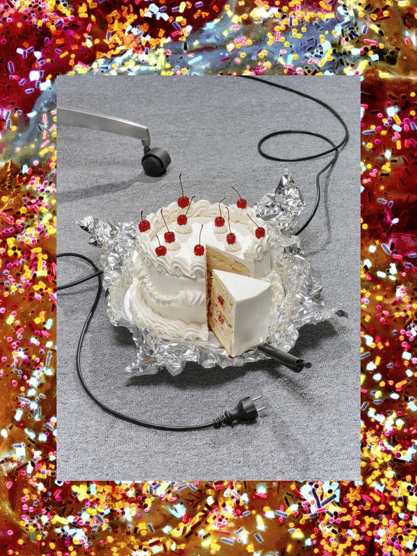 Das Bild zeigt einen weißen Kuchen mit roten Kirschen darauf, der in Scheiben geschnitten und auf ein Stück Alufolie gelegt wurde. Eine schwarze Schnur ist mit dem Kuchen verbunden, und im Hintergrund ist ebenfalls eine schwarze Schnur zu sehen. Der Kuchen steht vor einem Hintergrund mit einem bunten, glitzernden Muster.