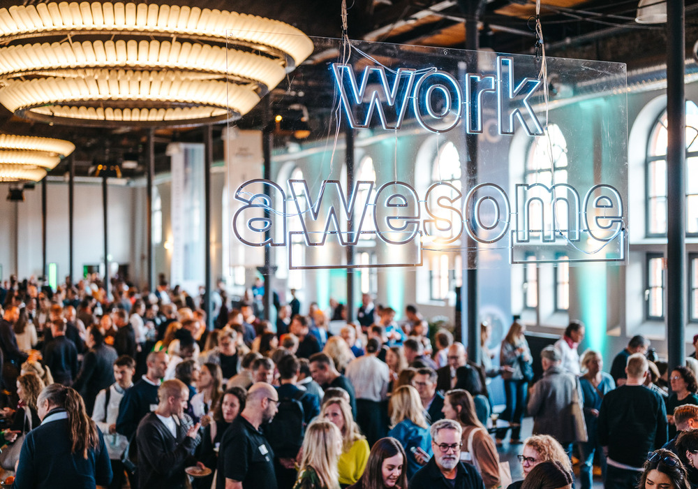 Das Bild zeigt eine große Menschenmenge, die sich in einem geräumigen Raum mit hohen Decken und großen Fenstern versammelt hat. Im Hintergrund ist eine auffällige Leuchtreklame mit der Aufschrift „work awesome“ zu sehen, die eine lebhafte und energiegeladene Atmosphäre schafft. Die Menschenmenge scheint verschiedenen Aktivitäten nachzugehen, was auf eine gesellschaftliche oder berufliche Veranstaltung hindeutet. Das Bild ist farbig und die Gesamtkomposition ist ausgewogen, wobei die Menschenmenge den größten Teil des Bildausschnitts ausfüllt.