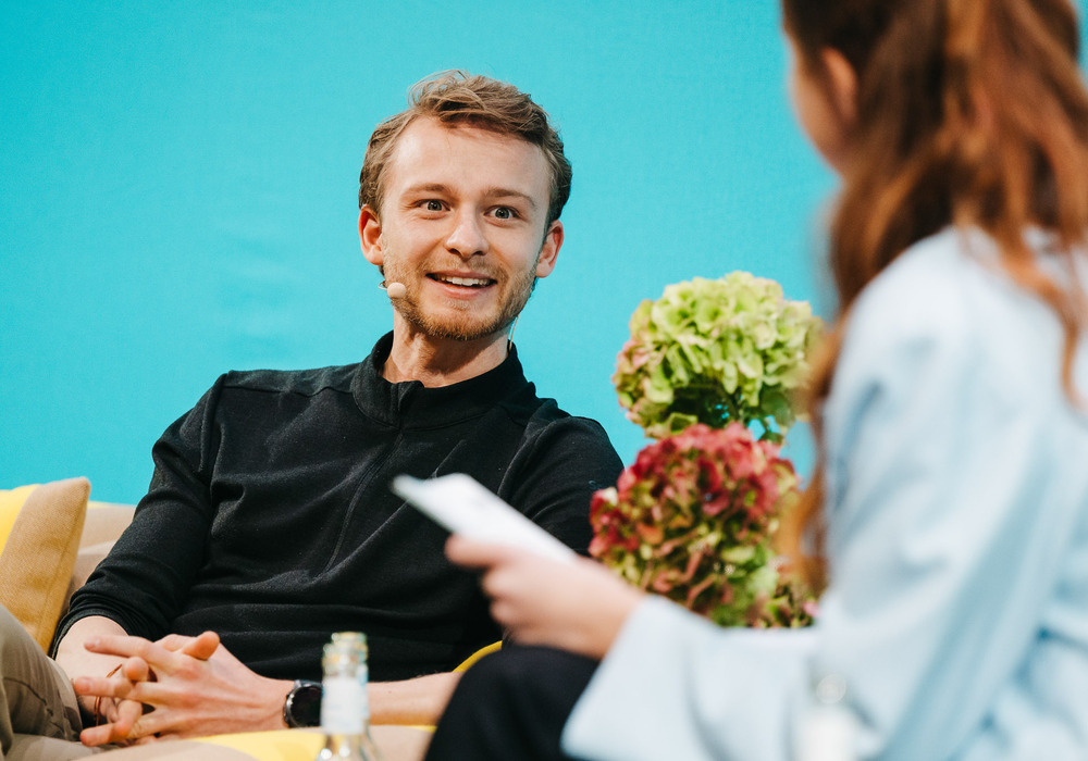 Das Bild zeigt einen Mann, der auf einer Couch sitzt, lächelt und sich mit einer Frau unterhält, die eine Fernbedienung in der Hand hält. Die Couch steht vor einem blauen Hintergrund, und in der Szene sind zwei Topfpflanzen zu sehen.