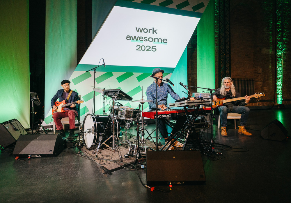 Das Bild zeigt eine Band, die auf einer Bühne auftritt, während im Hintergrund auf einer großen Leinwand die Worte „work awesome 2015” zu sehen sind. Die Bandmitglieder spielen verschiedene Instrumente, darunter Keyboard, Gitarre und Schlagzeug. Die Bühne ist gut ausgeleuchtet, und die Bandmitglieder stehen vor der Leinwand, wodurch eine mitreißende und energiegeladene Atmosphäre entsteht.