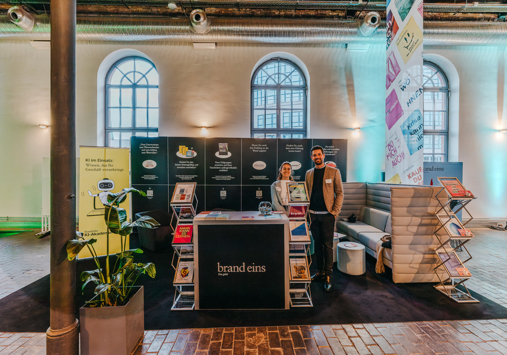 Das Bild zeigt einen Messestand, hinter dem ein Mann und eine Frau stehen. Der Stand ist in Schwarz-Weiß gehalten und präsentiert verschiedene Artikel, darunter Broschüren und Bücher. Außerdem sind einige Pflanzen und ein Topfgewächs zu sehen. Der Stand scheint für eine Messe oder Ausstellung aufgebaut worden zu sein, wobei der Schwerpunkt auf der Präsentation der angebotenen Produkte und Dienstleistungen liegt.