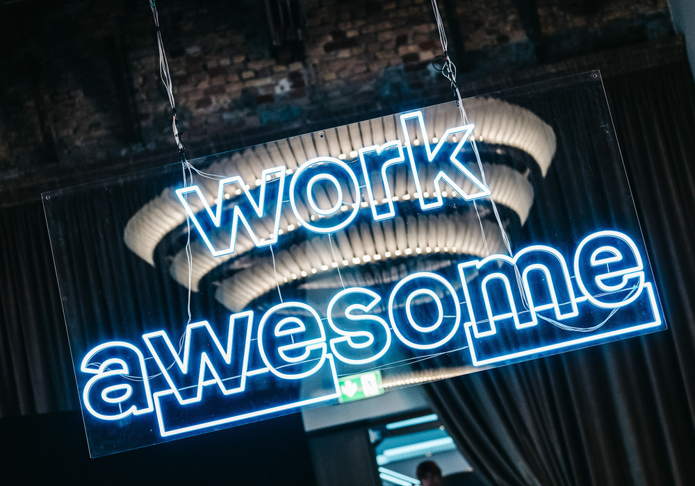 Das Bild zeigt eine Leuchtreklame mit der Aufschrift „work awesome“ in blauen Neonbuchstaben, die von der Decke hängt. Die Leuchtreklame ist beleuchtet und sorgt für eine helle und auffällige Darstellung. Der Hintergrund scheint ein dunkler Raum mit einer Backsteinwand zu sein, möglicherweise ein Restaurant oder eine Bar. Die Leuchtreklame steht im Mittelpunkt des Bildes, während die Hintergrundelemente den Kontext und die Tiefe der Szene vermitteln.