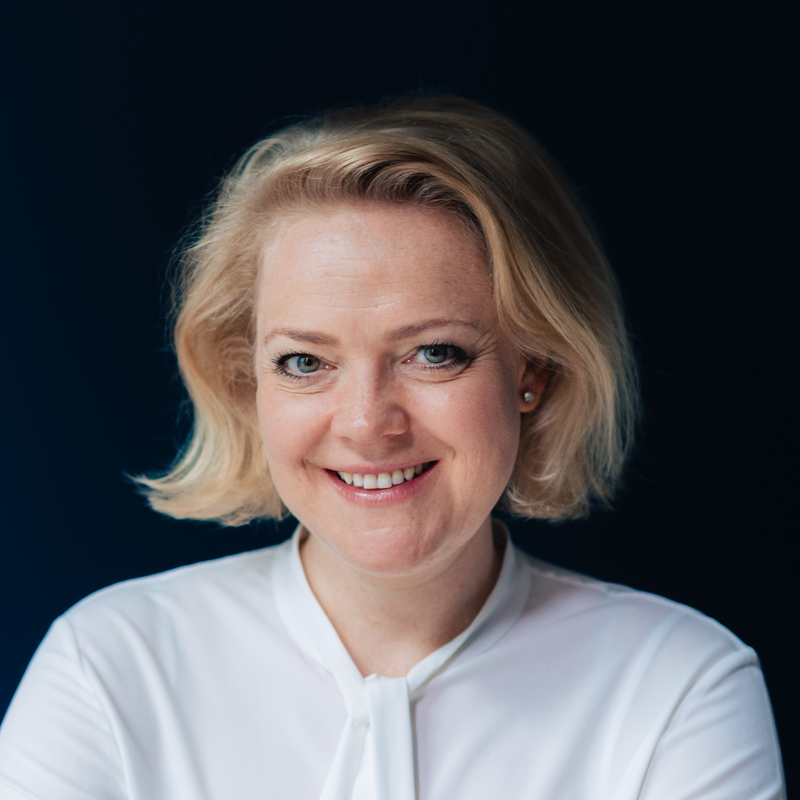 Das Bild zeigt eine lächelnde Frau mit blonden Haaren, die eine weiße Bluse trägt, vor einem dunklen Hintergrund.