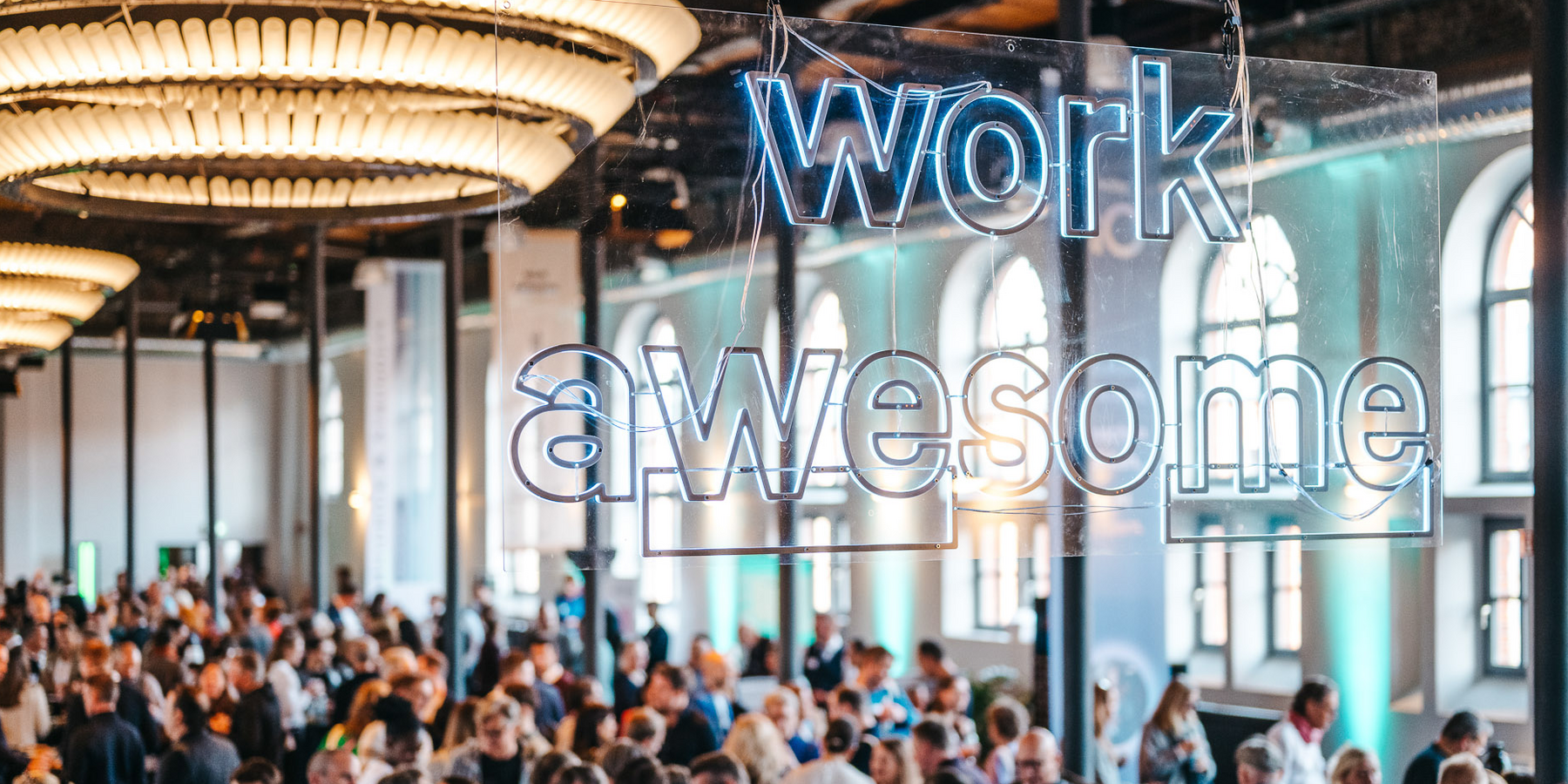 Das Bild zeigt eine große Menschenmenge, die sich in einem geräumigen Raum mit hohen Decken und großen Fenstern versammelt hat. Im Hintergrund ist eine auffällige Leuchtreklame mit der Aufschrift „work awesome“ in blauen Neonbuchstaben zu sehen. Die Reklame hängt von der Decke und bildet einen Blickfang in der Szene. Die allgemeine Atmosphäre wirkt lebhaft und energiegeladen, die Menschen sind mit verschiedenen Aktivitäten beschäftigt.