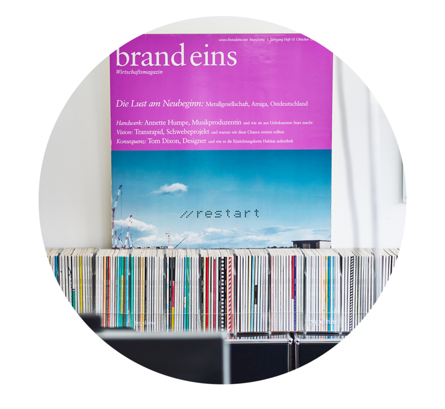 Das Bild zeigt eine Sammlung bunter Schallplatten, die ordentlich in einem Regal angeordnet sind, mit einem Magazin mit blauem Himmel und weißen Wolken, das an der Wand dahinter hängt. Das Magazin hat einen violetten Einband mit dem Titel „brandeins“ und den Aufdruck „Wirtschaftshafizmagazin“ und „Die Lust am Neuen: Metalgesellschaft, Amiga, Osterudland“. Auf dem Magazin ist auch der Text „resstart“ zu sehen.