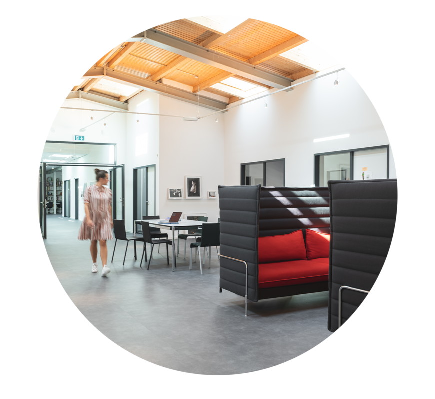 Das Bild zeigt einen modernen Büroraum, durch den eine Frau geht. Das Büro ist mit einer Kombination aus schwarzen und roten Möbeln ausgestattet, darunter ein rotes Sofa und schwarze Stühle. Außerdem sind mehrere Tische und Stühle im Raum verteilt, was auf eine kollaborative Arbeitsumgebung hindeutet. Das Gesamtdesign und die Raumaufteilung des Büros wirken gut durchdacht und funktional, wobei sowohl auf Ästhetik als auch auf Praktikabilität Wert gelegt wurde.