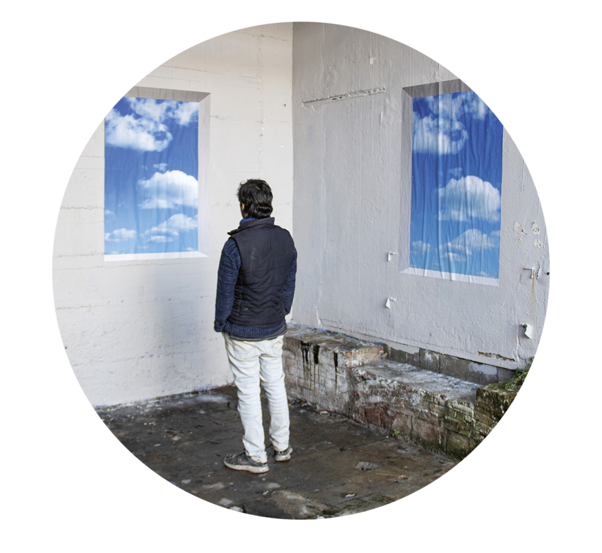 Das Bild zeigt eine Person, die in einem Raum mit zwei Fenstern steht, hinter denen jeweils ein blauer Himmel mit weißen Wolken zu sehen ist. Die Person steht mit dem Rücken zur Kamera und schaut aus den Fenstern hinaus. Der Raum wirkt heruntergekommen, mit Betonboden und Backsteinwänden. Die gesamte Szene vermittelt ein Gefühl von Einsamkeit und Nachdenklichkeit.