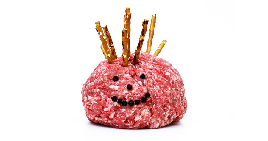 Das Bild zeigt einen Fleischbällchen mit einem Gesicht aus kleinen Brezelstangen und schwarzem Pfeffer. Der Fleischbällchen befindet sich auf einem weißen Hintergrund, wodurch ein optisch ansprechender Kontrast entsteht. Das Gesicht besteht aus mehreren Brezelstangen, die so angeordnet sind, dass sie ein Lächeln darstellen, was der Darstellung eine verspielte Note verleiht. Das Bild enthält keinen Text oder zusätzliche Objekte, und die relative Position der Objekte lässt auf eine Nahaufnahme des Fleischbällchens und seines Gesichts schließen.