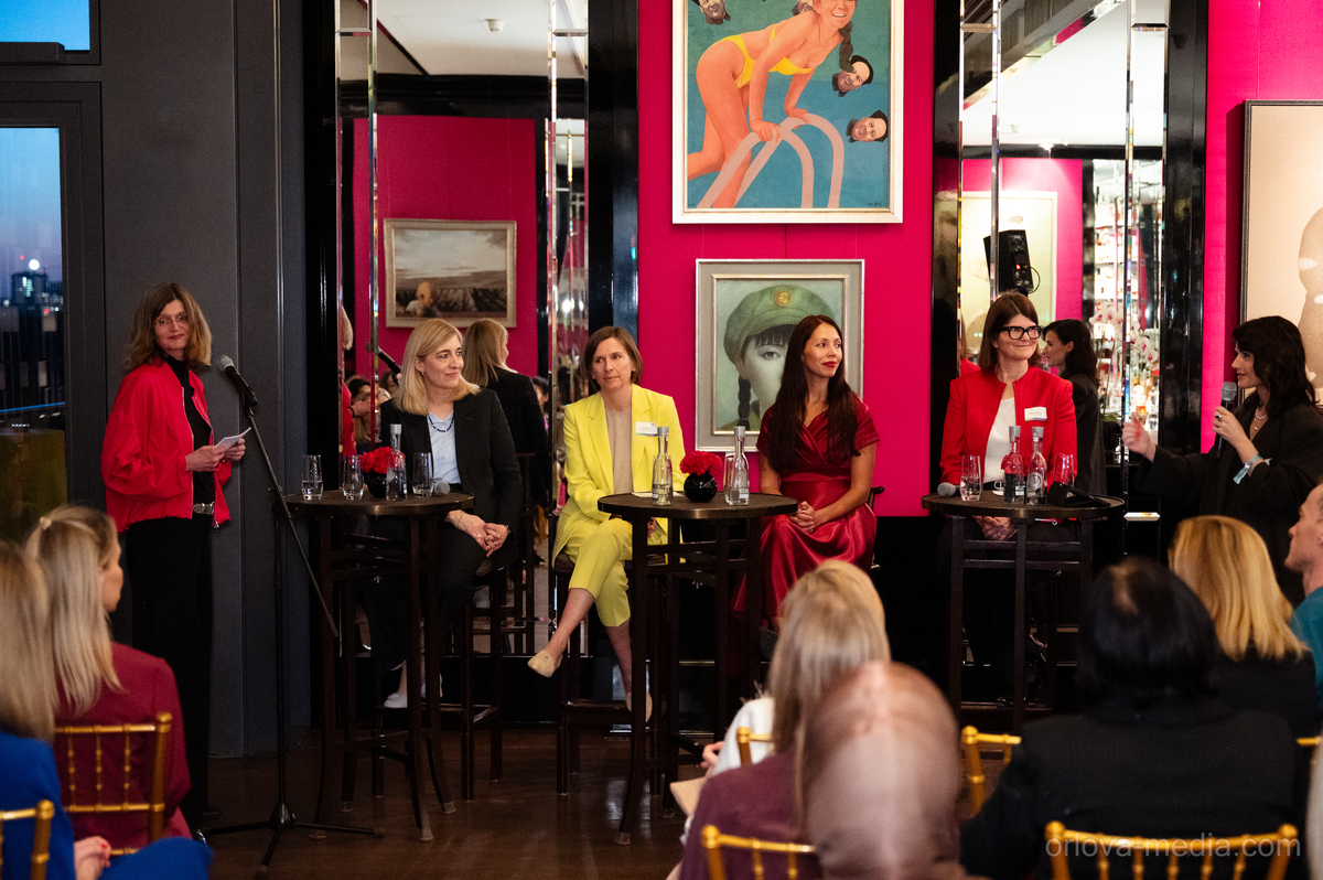 Das Bild zeigt eine Podiumsdiskussion, die in einem farbenfrohen Raum mit rosa Wänden stattfindet. Mehrere Personen sitzen an Tischen und unterhalten sich, während eine Person an einem Podium steht. Der Raum ist mit verschiedenen Kunstwerken und Spiegeln dekoriert, was eine künstlerische und lebendige Atmosphäre schafft. Das Publikum scheint der Podiumsdiskussion, die sich offenbar auf ein bestimmtes Thema konzentriert, aufmerksam zuzuhören.