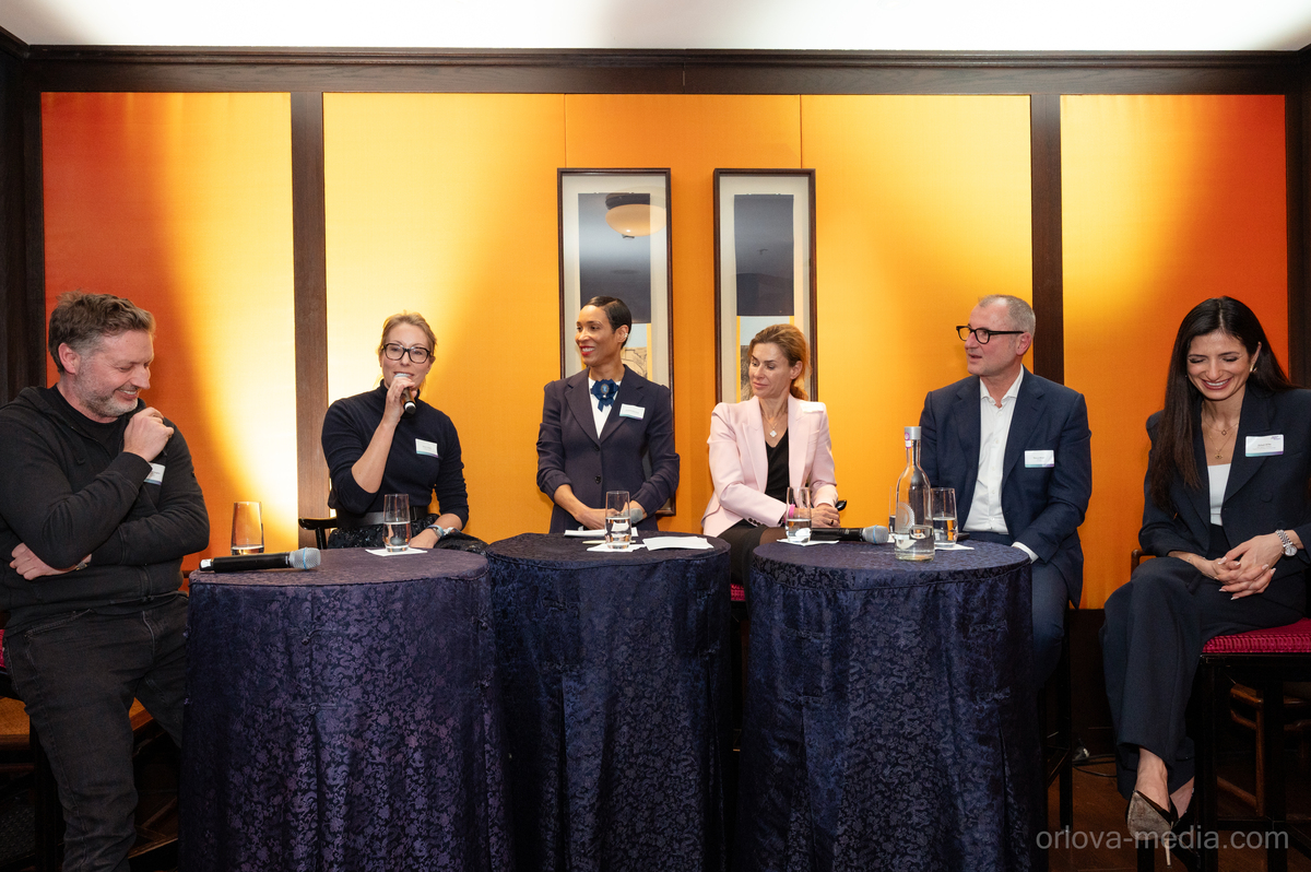 Das Bild zeigt eine Podiumsdiskussion mit sechs Personen, die an runden Tischen sitzen und jeweils ein Namensschild tragen. Die Teilnehmer sind in Business-Kleidung gekleidet, auf den Tischen stehen Gläser und Flaschen. Die Kulisse scheint eine formelle Veranstaltung oder Konferenz zu sein, mit einer warmen und einladenden Atmosphäre.