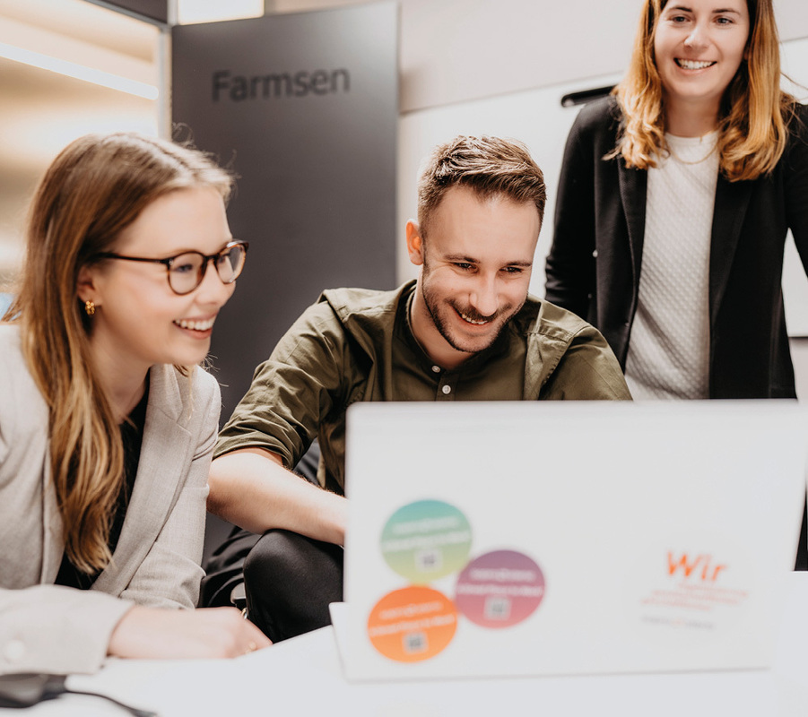 Das Bild zeigt drei Personen, zwei Frauen und einen Mann, die um einen Tisch sitzen und auf einen Laptop schauen. Auf dem Laptop befinden sich Aufkleber, und im Hintergrund ist ein Schild mit der Aufschrift „Farmsen“ zu sehen. Das Bild wird als barrierefrei für Menschen mit Behinderungen beschrieben und entspricht den EAA-Richtlinien.