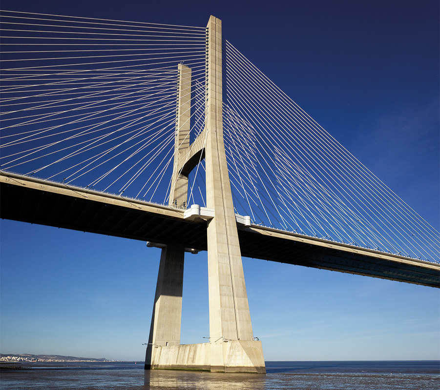 Das Bild zeigt eine große Hängebrücke mit mehreren Seilen und Pylonen, die sich über ein Gewässer spannt. Die Brücke steht vor einem strahlend blauen Himmel, und das Wasser darunter spiegelt die Konstruktion wider. Die Perspektive des Bildes ist von unten, mit Blick auf die beeindruckende Höhe und das Design der Brücke.