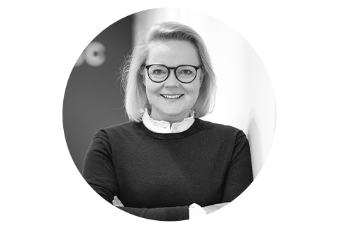 Das Bild ist ein Schwarz-Weiß-Porträt einer Frau mit Brille, die vor einer Wand steht. Sie trägt einen Pullover und einen Kragen, was ihr ein professionelles Aussehen verleiht. Die Frau lächelt und hat die Arme vor sich verschränkt. Das Bild ist von einem Rahmen umgeben, und es ist kein Text vorhanden.