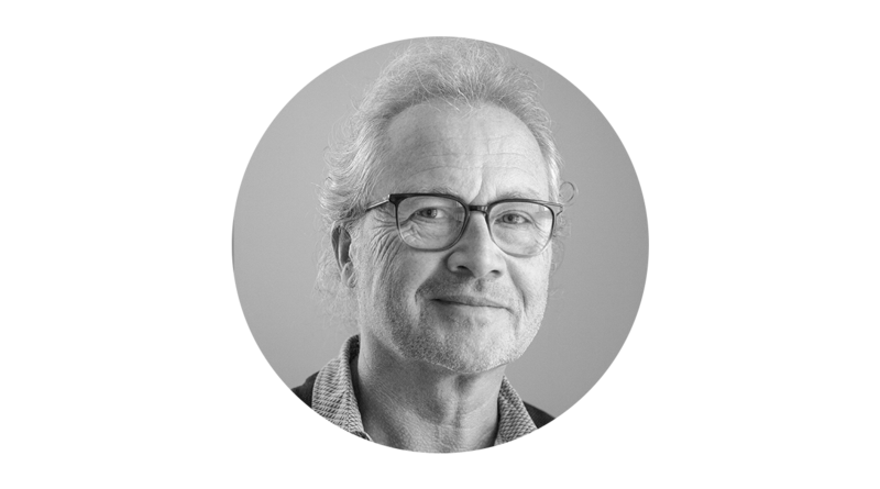 Das Bild zeigt ein Schwarz-Weiß-Porträt eines älteren Mannes mit Brille, der mit einem leichten Lächeln direkt in die Kamera blickt. Der Mann trägt ein Hemd mit Kragen, der Hintergrund ist schlicht weiß. Das Bild ist quadratisch, und es ist kein Text zu sehen.