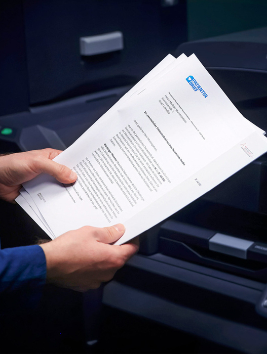 Eine Person hält einen Stapel weißer Blätter mit blauem Text in der Hand, bei denen es sich offenbar um ein Dokument oder einen Bericht handelt. Die Person steht vor einem Drucker, was darauf hindeutet, dass die Dokumente gerade gedruckt oder bearbeitet werden. Das Bild ist in Farbe, und es ist kein Text zu sehen, der nicht klar lesbar wäre. Im Mittelpunkt der Szene scheint der Druckvorgang zu stehen, wobei die Person vermutlich damit beschäftigt ist, die Dokumente zu prüfen oder für die Verteilung vorzubereiten.