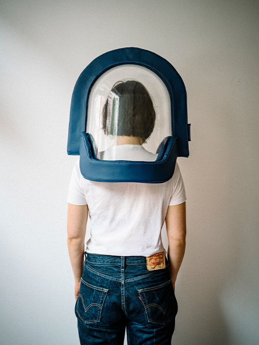 Das Bild zeigt eine Person, die vor einer weißen Wand steht und ein weißes T-Shirt sowie eine blaue Jeans trägt. Sie trägt einen blauen Helm mit einem durchsichtigen Visier, das den Kopf bedeckt und das Gesicht verdeckt. Der Helm scheint für einen bestimmten Zweck konzipiert zu sein, möglicherweise im Zusammenhang mit der Luftfahrt oder der Sicherheit.