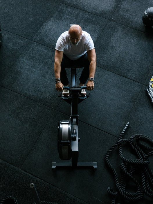 Das Bild zeigt einen Mann, der in einem Fitnessstudio auf einem stationären Heimtrainer sitzt. Der Mann trägt ein weißes T-Shirt und scheint sich ganz auf sein Training zu konzentrieren. Der Heimtrainer steht in der Bildmitte, und auf dem Boden rechts im Bild ist ein Seil zu sehen. Die gesamte Szene vermittelt ein Gefühl von Fitness und Engagement für körperliche Betätigung.
