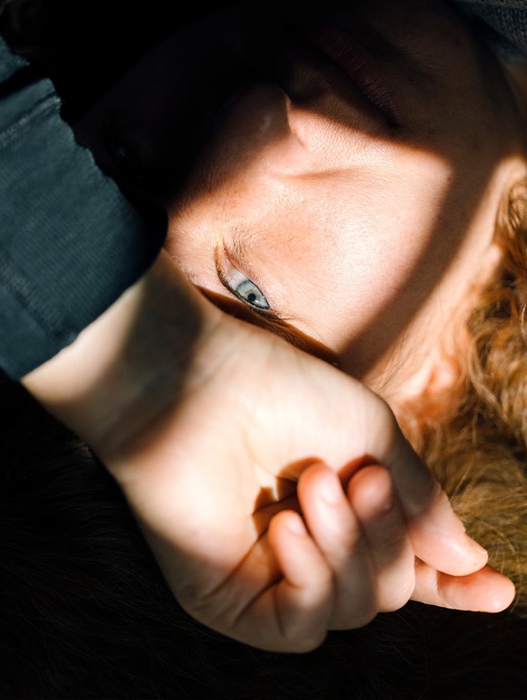 Das Bild zeigt eine Nahaufnahme des Gesichts einer Person, deren Augen sanft geschlossen sind und deren Hand auf dem Kinn ruht. Das Gesicht der Person wird teilweise vom Schatten ihrer Hand verdeckt, wodurch ein Gefühl von Tiefe entsteht und der Blick auf die Augen gelenkt wird. Der Hintergrund ist dunkel, was das Gesicht der Person und das Wechselspiel von Licht und Schatten noch stärker hervorhebt.