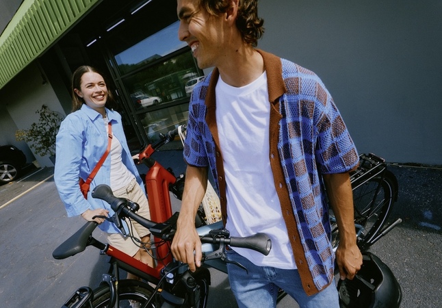 Ein Mann und eine Frau stehen lächelnd neben einem abgestellten Fahrrad; es sieht so aus, als befänden sie sich auf einem Parkplatz. Der Mann hält den Lenker des Fahrrads fest, während die Frau hinter ihm steht.