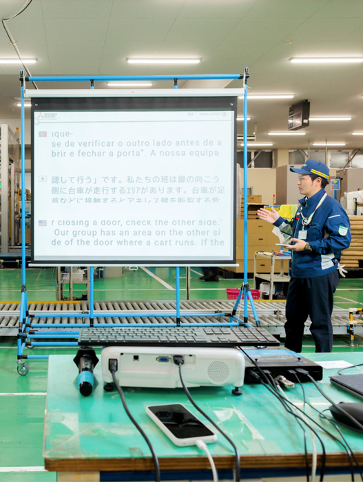 Das Bild zeigt einen Mann in blauer Uniform, der vor einem großen Bildschirm steht, auf dem Text in portugiesischer Sprache zu sehen ist. Der Bildschirm ist auf einem blauen Ständer befestigt, und auf dem Tisch vor dem Mann sind verschiedene elektronische Geräte und Kabel zu sehen. Die Szene scheint in einer Werkstatt oder einer Fabrikumgebung zu spielen, wobei der Mann dem Publikum möglicherweise etwas vorstellt oder erklärt.