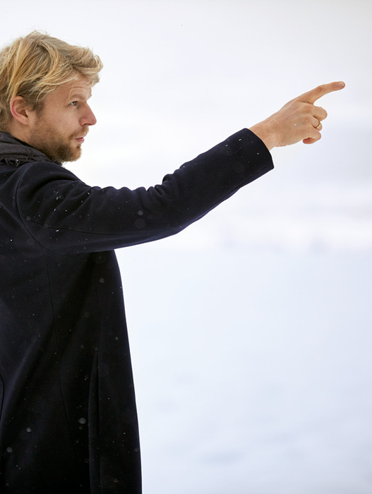 Das Bild zeigt einen Mann in einem schwarzen Mantel, der im Freien steht und mit dem Finger auf die rechte Seite des Bildausschnitts zeigt. Der Hintergrund ist weiß und verschwommen, was möglicherweise auf Schnee oder einen bewölkten Himmel hindeutet. Der Mann scheint sich in einer verschneiten Umgebung zu befinden, möglicherweise in einer winterlichen Kulisse.